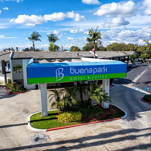 The image shows the exterior of Buena Park Hotel & Suites, featuring a prominent sign, palm trees, and a clear, sunny sky in the background.