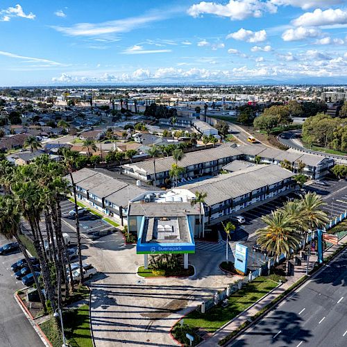 Aerial view of a sprawling cityscape featuring a hotel complex surrounded by roads, palm trees, greenery, and distant buildings under a partly cloudy sky.