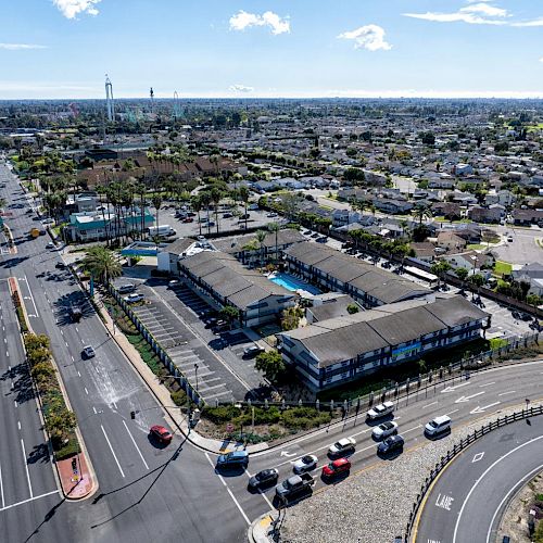 An aerial view of a suburban area with roads, cars, buildings, and houses sprawling in the distance under a blue sky with scattered clouds.