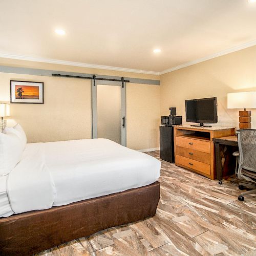 A modern hotel room with a double bed, wooden desk, flat-screen TV, chair, and sliding door, featuring warm lighting and stylish decor.