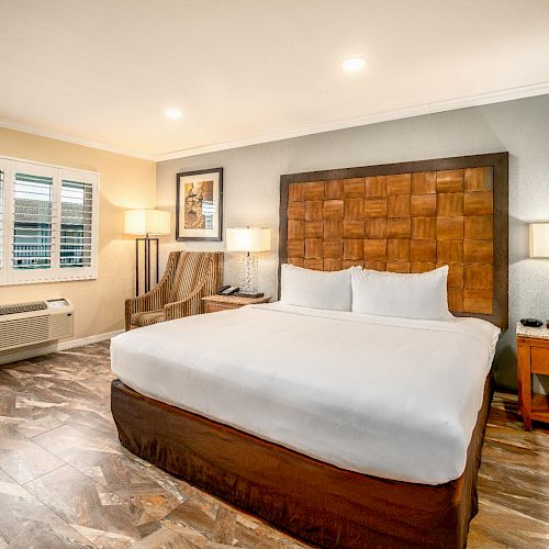 The image shows a modern hotel room with a large bed, two bedside tables, a chair, a lamp, and a window with shutters.