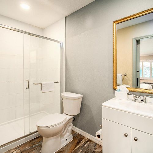 A modern bathroom with a glass-enclosed shower, toilet, a vanity with a sink, toiletries, and a large mirror in a gold frame.