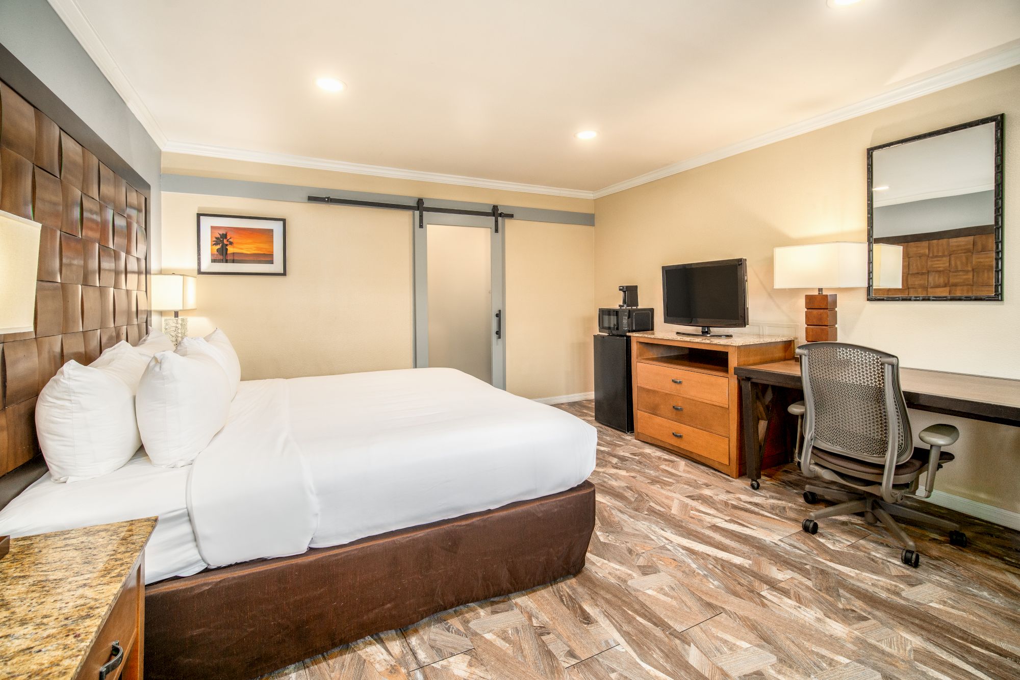 A neatly arranged hotel room with a king-size bed, wall art, desk and chair, TV, and a sliding glass door. The room has a warm and inviting ambiance.