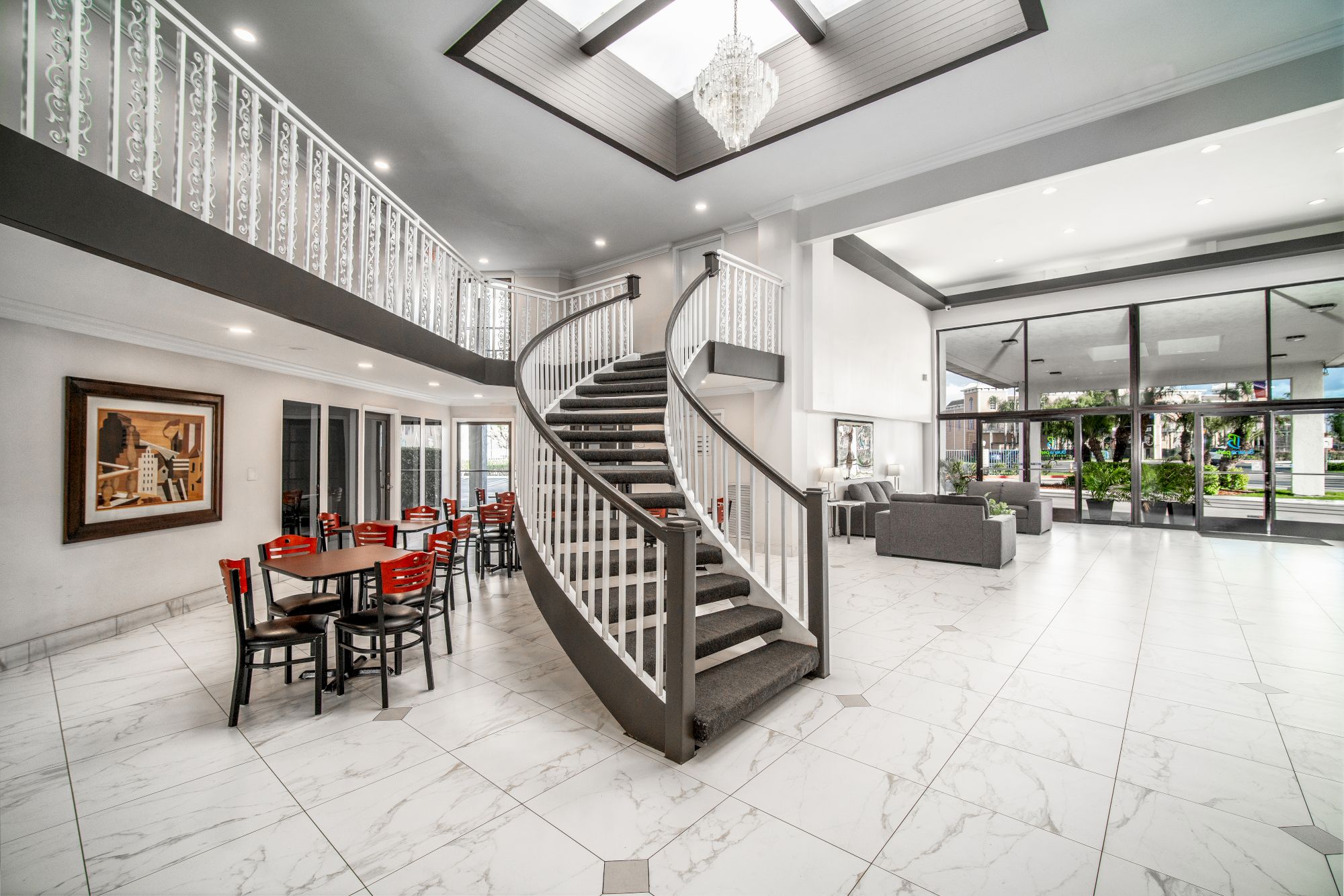 This image shows a spacious, modern lobby with a grand curved staircase, white interiors, artwork, red chairs at a table, and large windows for natural light.