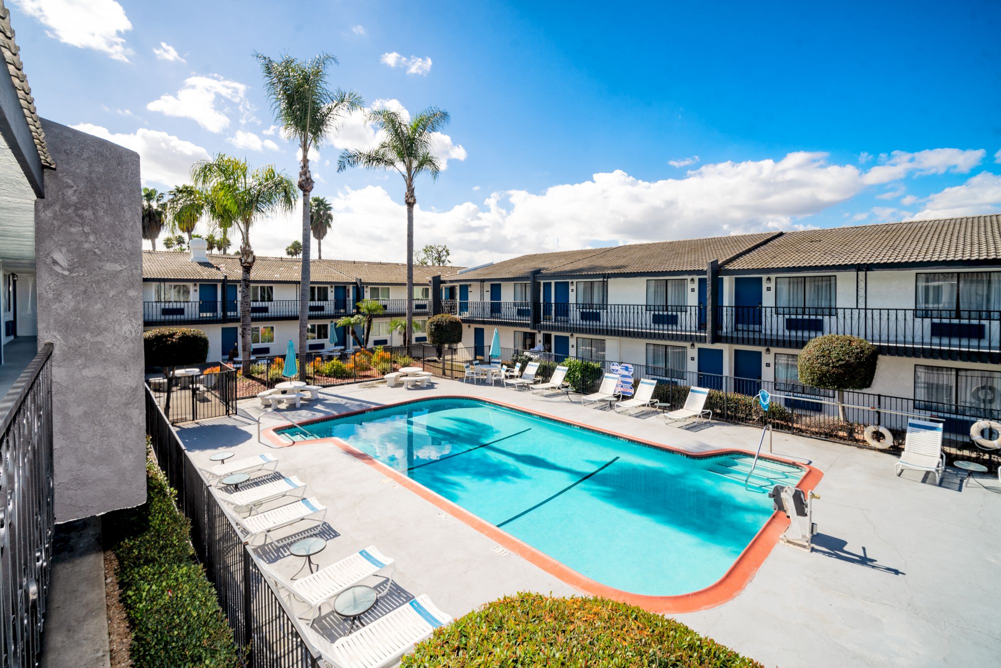 This image shows an outdoor swimming pool surrounded by lounge chairs in the center of a motel-style building complex with palm trees and a blue sky.