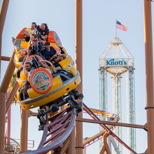 People riding a roller coaster at Knott's Berry Farm with the park's tower and an American flag in the background.