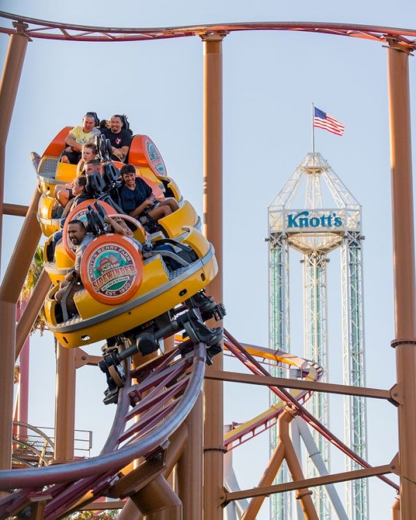 People riding a roller coaster at Knott's Berry Farm with the park's tower and an American flag in the background.