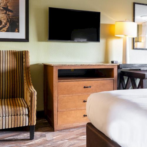 The image shows a hotel room with a striped armchair, a dresser with a TV, a bed, a desk, a chair, a lamp, and framed art on the wall.