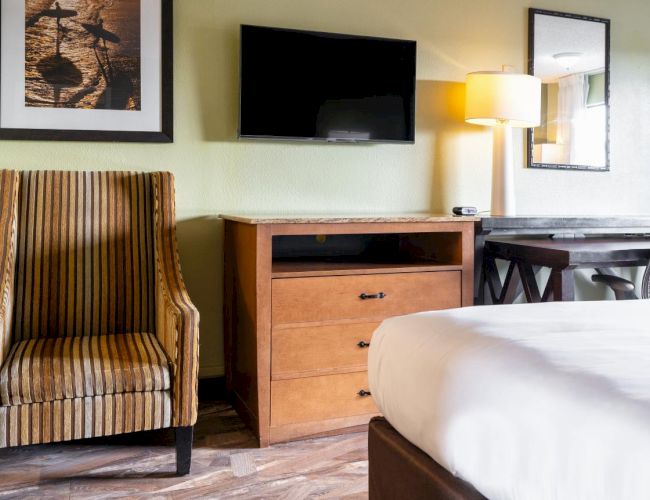 The image shows a hotel room with a striped armchair, a dresser with a TV, a bed, a desk, a chair, a lamp, and framed art on the wall.