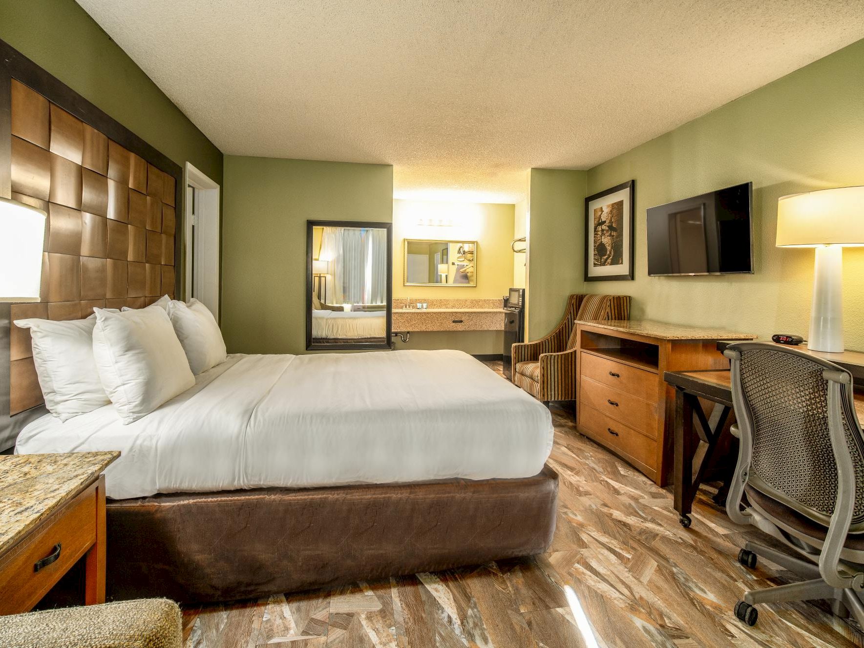 A hotel room with a king-sized bed, desk, TV, and chair.