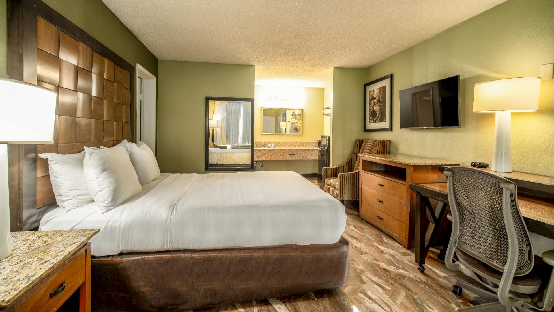 A hotel room with a king-sized bed, desk, chair, TV, lamps, and a bathroom area visible. The decor is modern with green and brown tones.