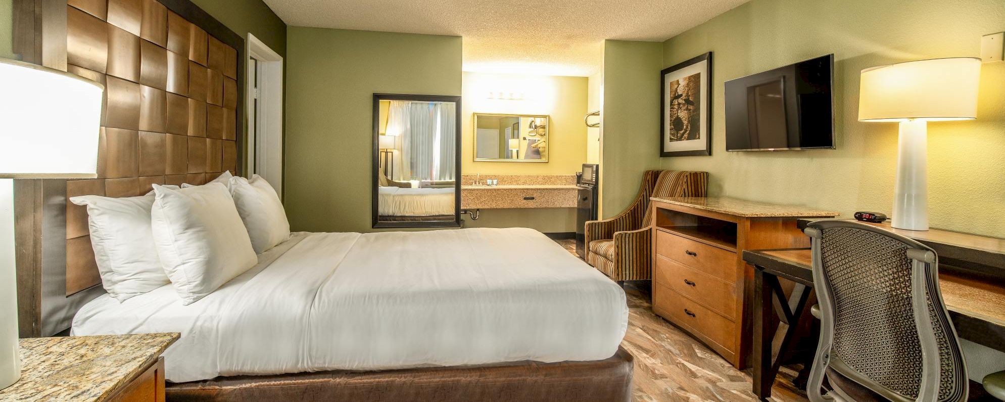 A wide view of a hotel room with a king-size bed, desk, TV, and bathroom mirror.