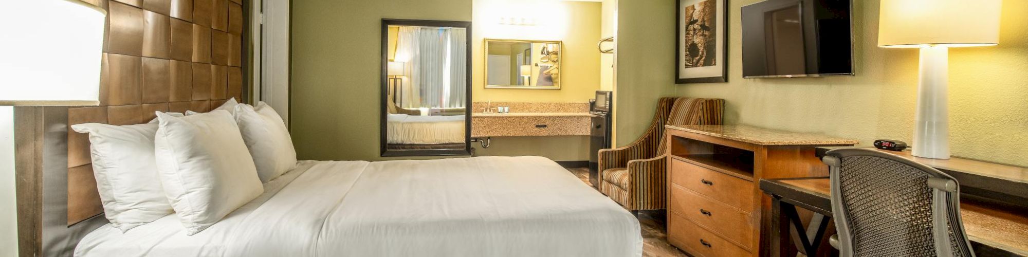 A hotel room with a king-sized bed, desk, chair, TV, lamps, and a bathroom area visible. The decor is modern with green and brown tones.