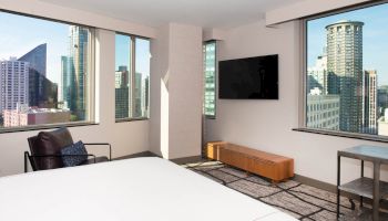 A modern hotel room features a bed, a wall-mounted TV, and large windows with a cityscape view, including skyscrapers and high-rise buildings.