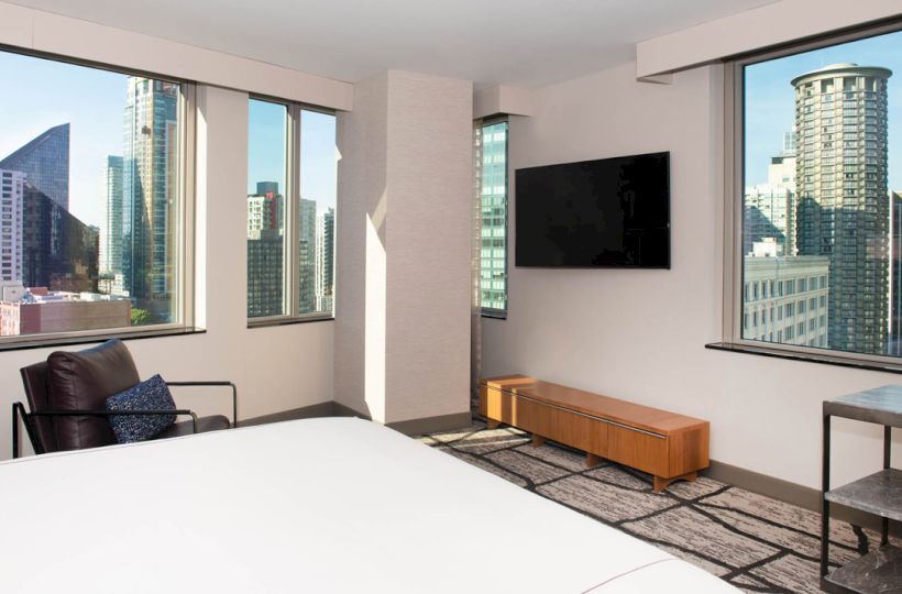 A modern hotel room features a bed, a wall-mounted TV, and large windows with a cityscape view, including skyscrapers and high-rise buildings.