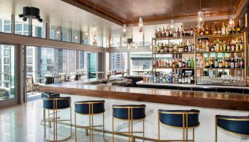 A modern, upscale bar with a long wooden counter, five bar stools, hanging lights, and a well-stocked liquor shelf, overlooking a cityscape.