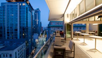 The image shows a modern cityscape at dusk with high-rise buildings and a rooftop terrace featuring seating areas and tables.