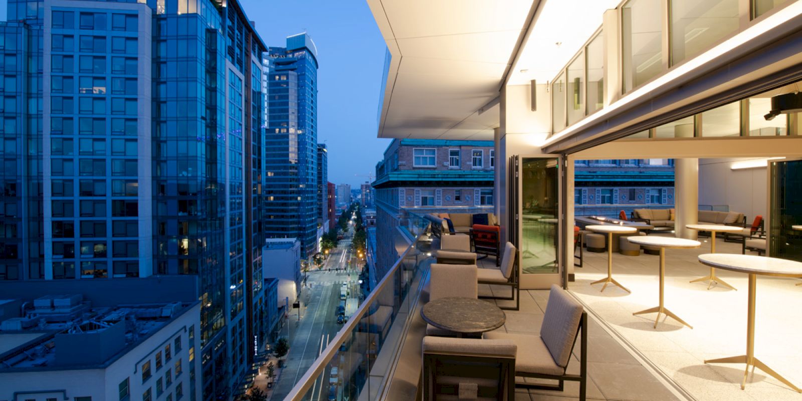 A modern balcony with sleek furniture overlooks a city street and high-rise buildings during dusk, with lights illuminating the urban scene.
