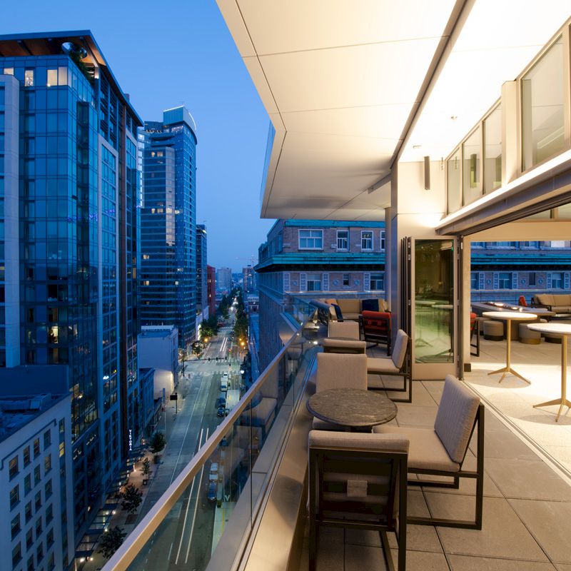 A rooftop terrace at dusk with tables and chairs, overlooking a cityscape of modern high-rise buildings and a busy street below.