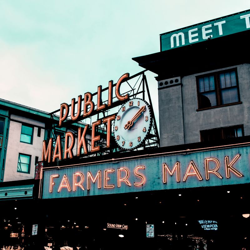 This image shows the iconic signs of a "Public Market" and "Farmers Market" at a marketplace, along with a clock and surrounding buildings.