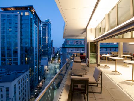 A modern city-setting balcony at twilight features outdoor seating and tables, overlooking tall buildings and a street below.