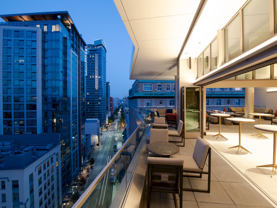 A modern balcony overlooking an urban city street at dusk. There are tables and chairs set up on the balcony with buildings in the background.