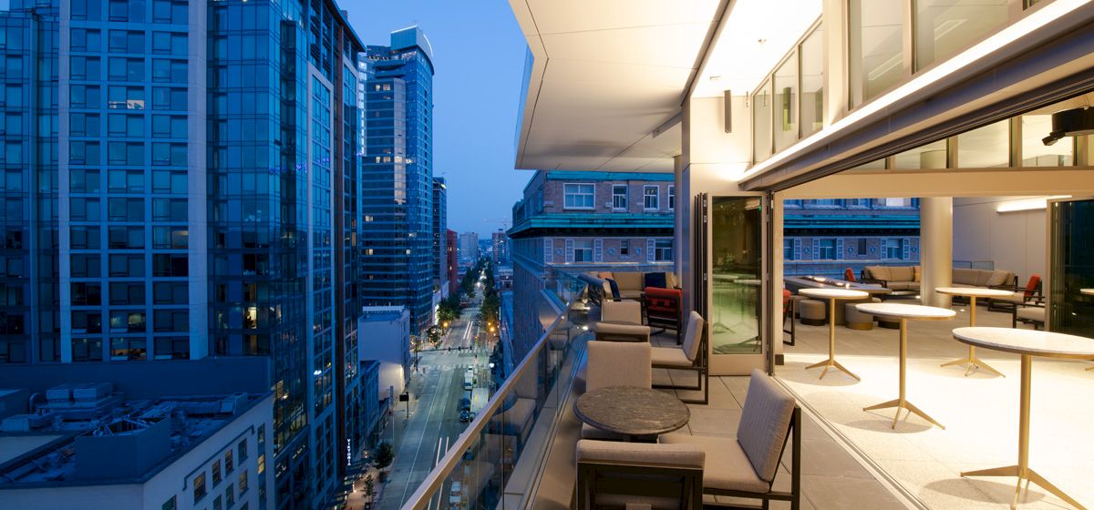 A modern balcony overlooking an urban city street at dusk. There are tables and chairs set up on the balcony with buildings in the background.