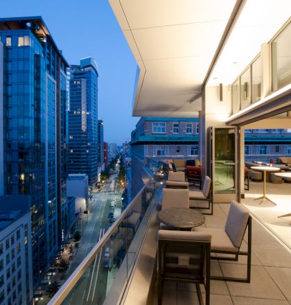 A modern cityscape at dusk with high-rise buildings and a luxurious open balcony with seating and tables overlooking the urban streets below.