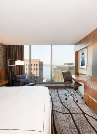 A modern hotel room features a large bed, a wall-mounted TV, a desk, and a chair, with a window view of buildings and the waterfront.