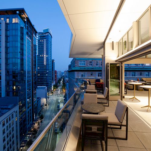 The image shows a modern balcony with chairs and tables overlooking a cityscape with tall buildings and streetlights at dusk.