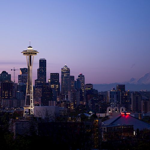 The image shows the Seattle skyline at dusk, featuring the Space Needle prominently and Mount Rainier in the background against a twilight sky.