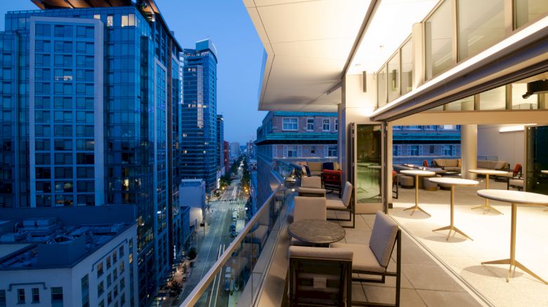 A modern balcony view of a cityscape at dusk, featuring outdoor seating and tables. Indoor area with more seating visible through a large glass wall.