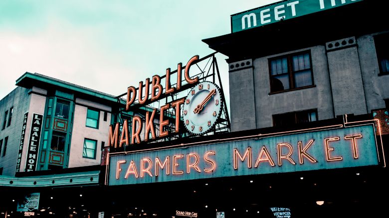 The image shows the exterior of a public farmers market with neon signs reading "Public Market" and "Farmers Market." The clock adds a vintage feel.