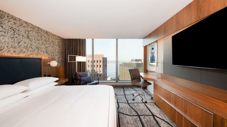 A modern hotel room with a large bed, desk, chair, and a wall-mounted TV, featuring floor-to-ceiling windows with a cityscape view.