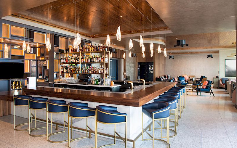 A sleek and modern bar with hanging lights, a well-stocked liquor shelf, blue barstools, and seating areas in the background.