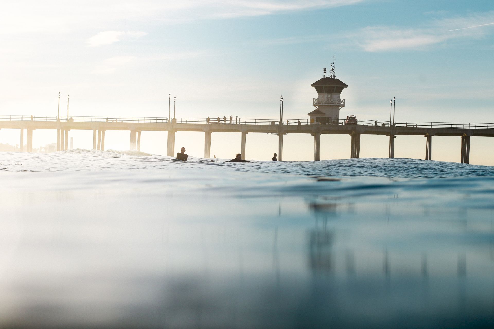 A serene image shows a pier extending into the ocean with a few surfers in the water, and a building and people on the pier in the background.
